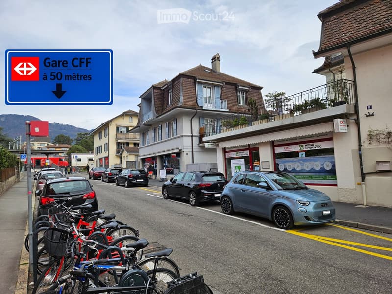 Street view showing parked cars, bicycles, and buildings with signage for a train station nearby.