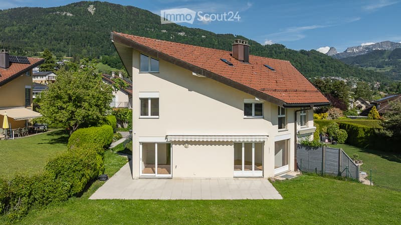 Two-story house with a red-tiled roof, large windows, and a well-maintained garden, set against a scenic mountain backdrop.