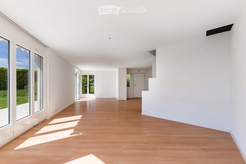 Empty hallway with wooden flooring, white walls, large windows, and a staircase leading to another level.