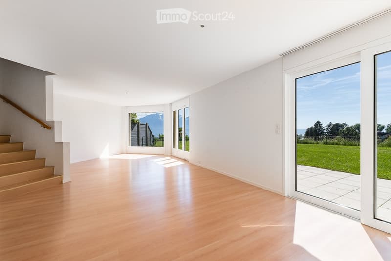 Bright, empty room with wooden flooring, white walls, large windows, and a staircase leading upwards.