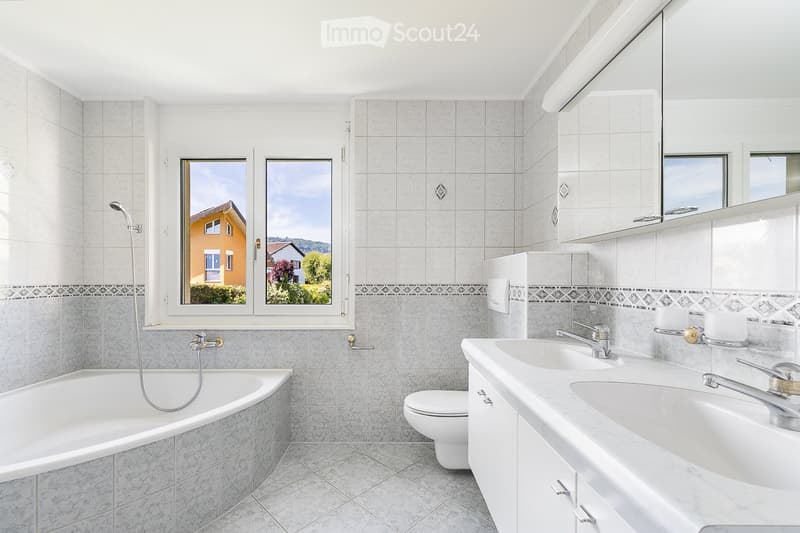 Modern bathroom with a bathtub, toilet, double sink, and large windows offering a view of the exterior.