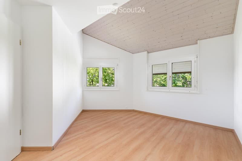 Empty room with white walls, light wooden flooring, and large windows with white frames. The ceiling is made of light wood panels.