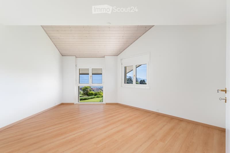 Empty room with white walls, wooden floor, and large windows offering a view of greenery and mountains.