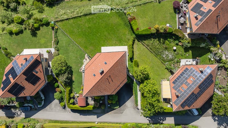 Aerial view of a residential area with multiple houses featuring red-tiled roofs and solar panels, surrounded by well-maintained green lawns and gardens.