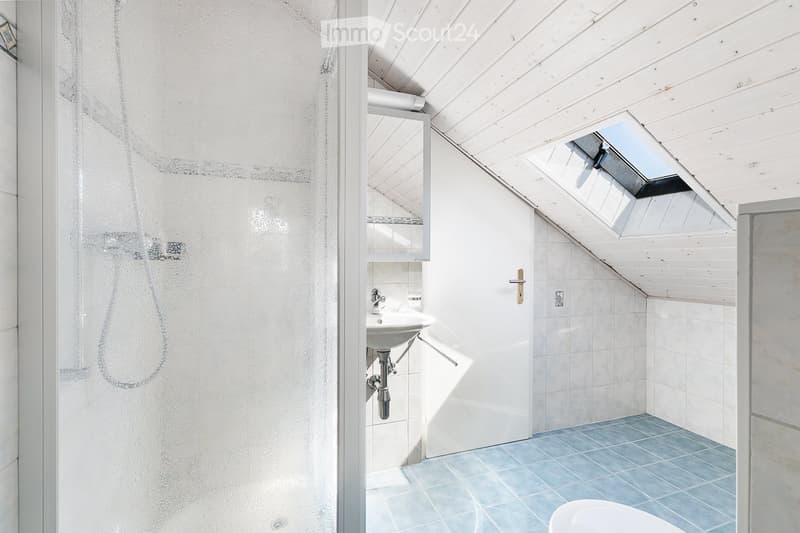Modern bathroom with white tiled walls, blue tiled floor, a sink with a mirror, and a toilet. Features a skylight window and a glass shower enclosure.