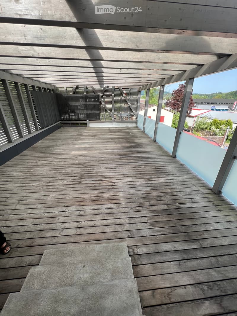 A wooden deck with a concrete staircase leading to it, surrounded by a metal railing. The deck is empty and has a view of the outside area.