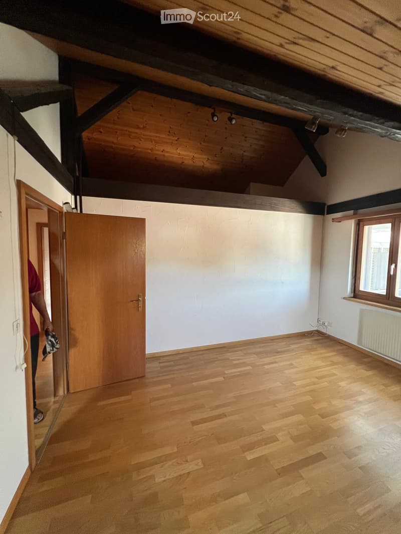 Empty room with wooden floor, white walls, wooden ceiling, and a window. There is a door on the left and a radiator on the right wall.
