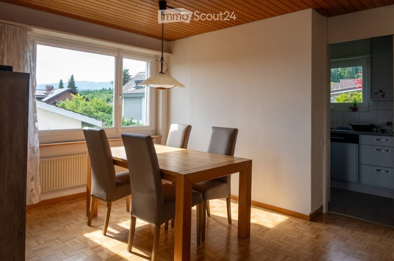 Bright dining area with a wooden table and four chairs, adjacent to a modern kitchen with cabinets and a dishwasher. Large windows offer a scenic view of the surrounding landscape.
