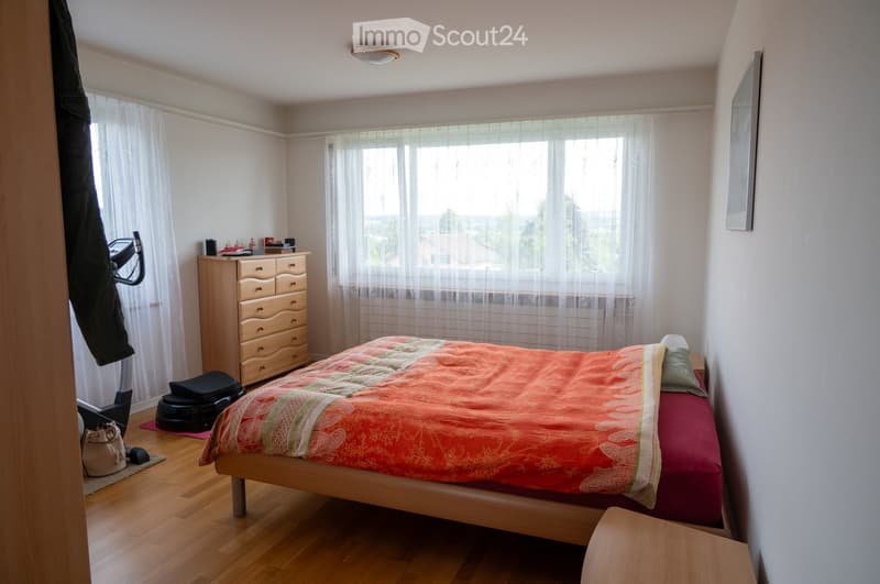Well-lit bedroom with wooden flooring, large window with sheer curtains, bed with colorful bedding, dresser, nightstand, exercise bike, and personal items near the door.