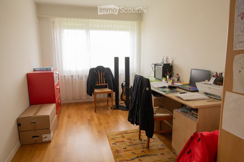 A furnished room with wooden flooring, a large window with white curtains, a desk with a laptop, printer, and various office supplies, a guitar, a speaker, a chair with a jacket draped over it, and a colorful rug.