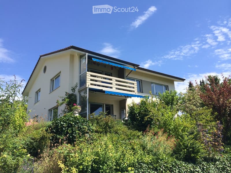 Two-story house with a balcony, surrounded by lush greenery and flowers.