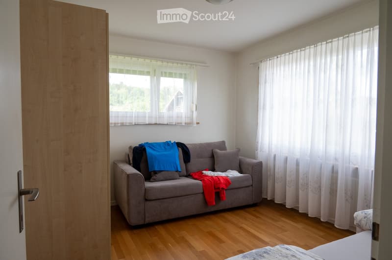 A small, furnished living room with a grey sofa, pillows, and clothes draped over it. The room has wooden flooring, white walls, and large windows with white curtains.