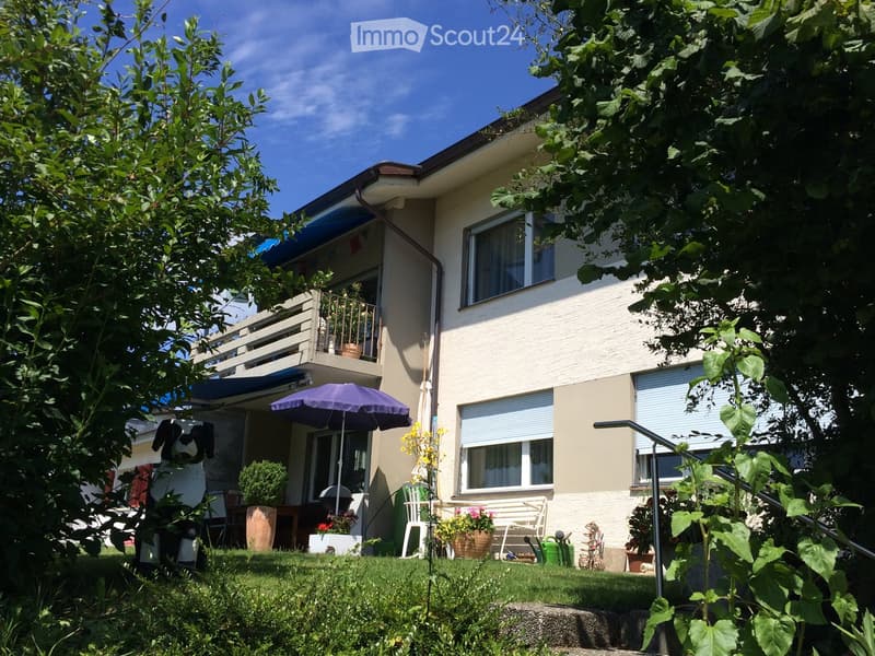 A two-story house with a balcony, surrounded by a well-maintained garden with various plants, a bench, and outdoor furniture.