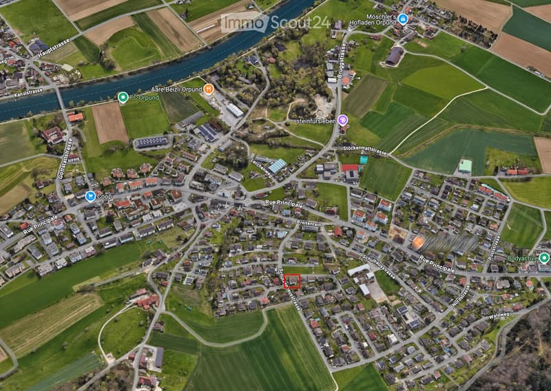 Aerial view of a residential area with houses, streets, and surrounding green fields. The area includes a mix of residential buildings, roads, and agricultural land.
