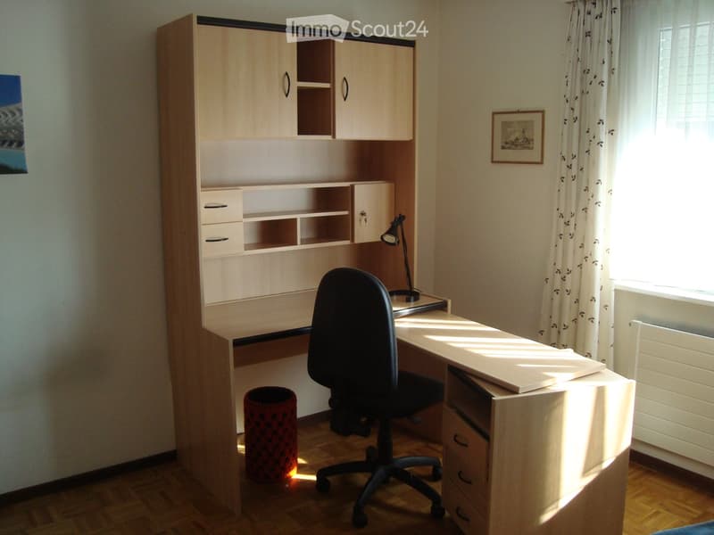 Light wood desk, 2 drawers on the left, 2 shelves above, 1 drawers on the right side, black office chair, black desk lamp, open photo frame in the right top corner, radiator below window, window with white curtain