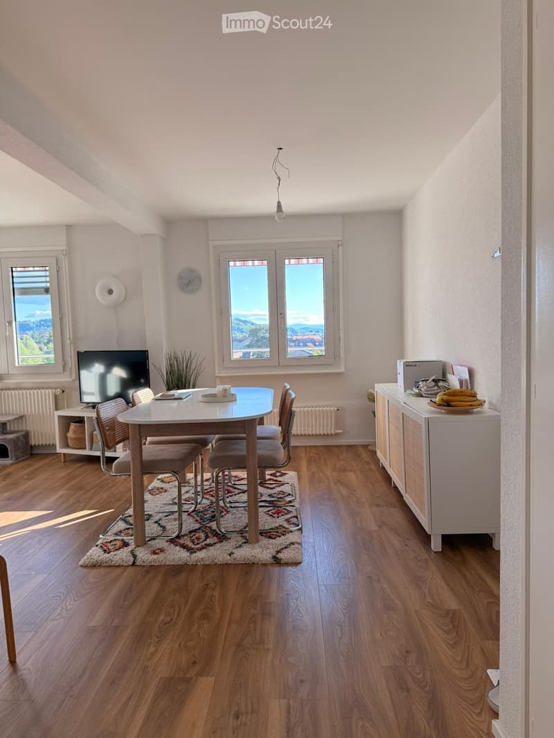 Bright dining area with wooden flooring, large windows offering a scenic view, a round dining table with four chairs, a television on a stand, a cabinet with a fruit bowl, and a small plant in the corner.