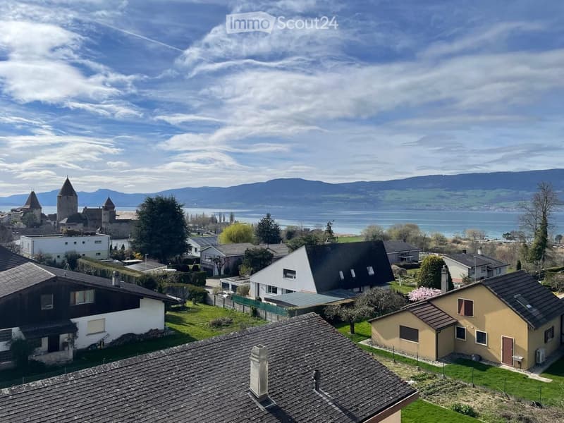 Aerial view of a residential area with houses, gardens, and a distant lake and mountains in the background.