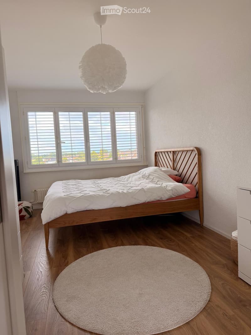 Bright bedroom with wooden flooring, a white pendant light, a round rug, a wooden bed with white bedding, and a round window with blinds overlooking a scenic view.