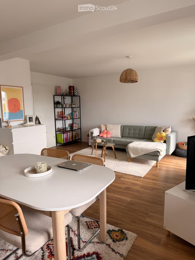 A modern living room with a grey sofa, wooden coffee table, white dining table, and a hanging wicker lamp. The room features wooden flooring, colorful rugs, and decorative shelves.