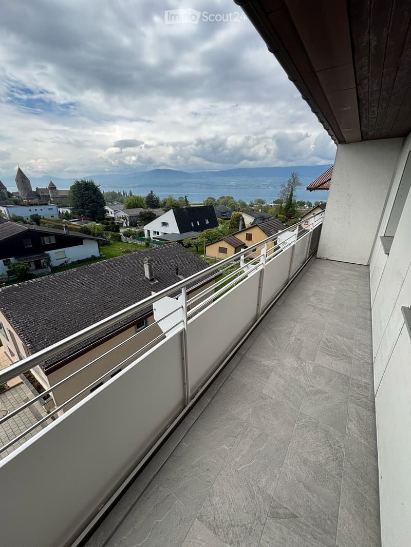 Balcony with a modern metal railing, tiled floor, overlooking a residential area with houses, trees, and a distant lake and mountains.