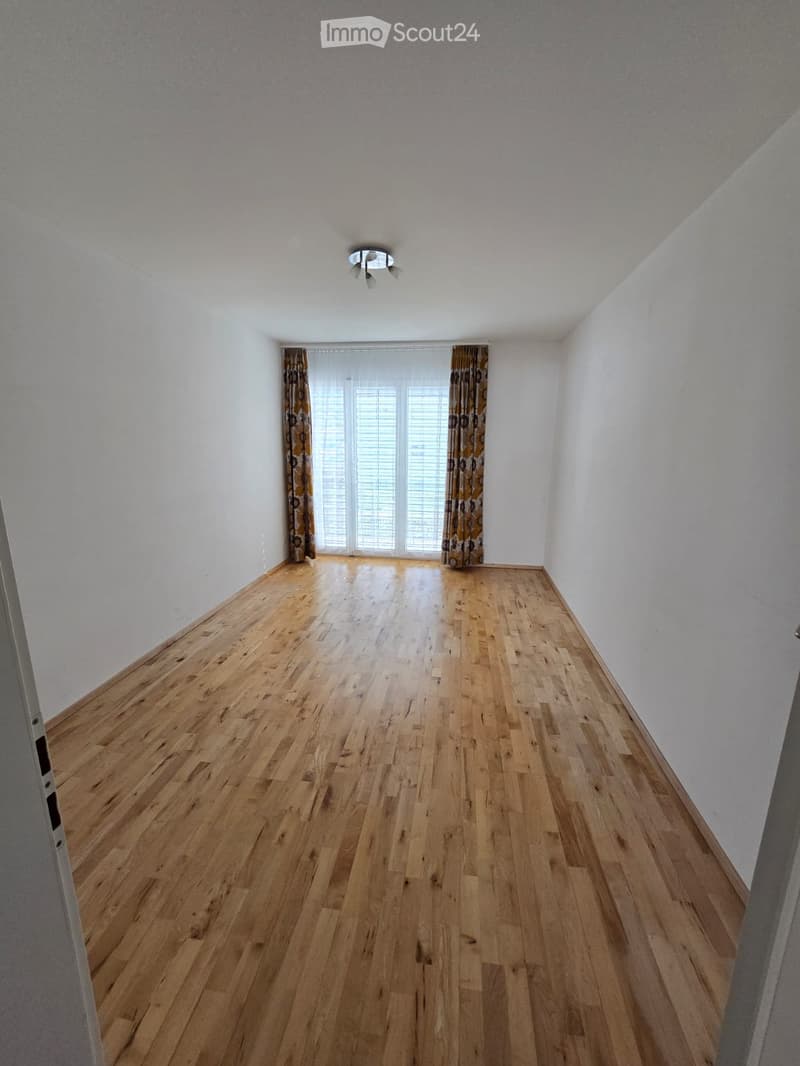 Empty room with wooden flooring, white walls, and large windows with blinds and curtains.