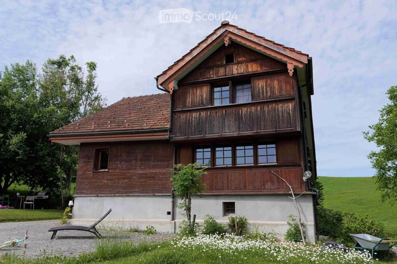 Traditional two-story wooden house with a red-tiled roof, wooden balcony, and brick lower section. Surrounded by a garden with white flowers, a lounge chair, and a wheelbarrow.
