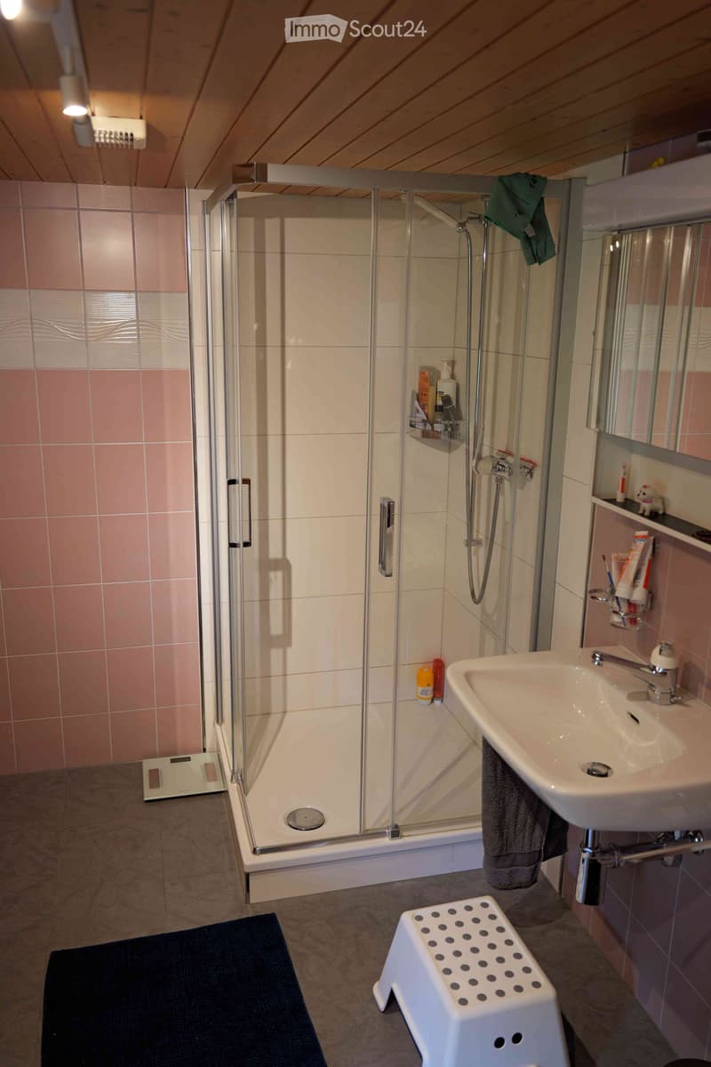 Modern bathroom with a glass-enclosed shower, a white sink with a chrome faucet, a towel hanging on the side, a small white stool with black dots, and a dark blue bath mat on the floor.