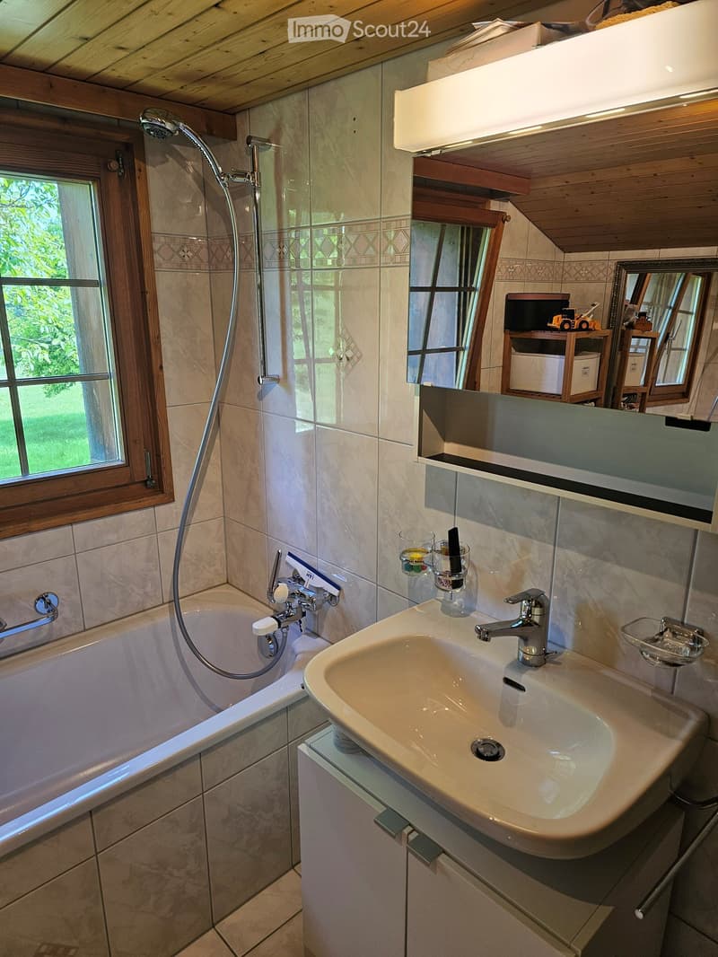 Bathroom with a bathtub, sink, mirror, and shower hose. Wooden ceiling and tiled walls.