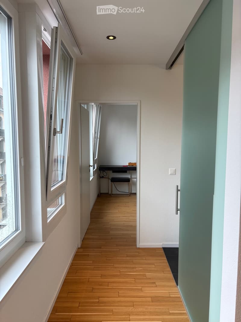 A narrow hallway with wooden flooring, white walls, and large windows on one side. A small room with a desk and chair is visible at the end.