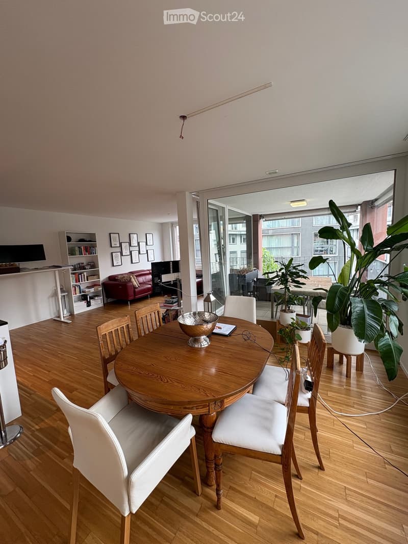 Dining area with a round wooden table, white chairs, a red sofa, bookshelf, and large windows leading to a balcony with plants.