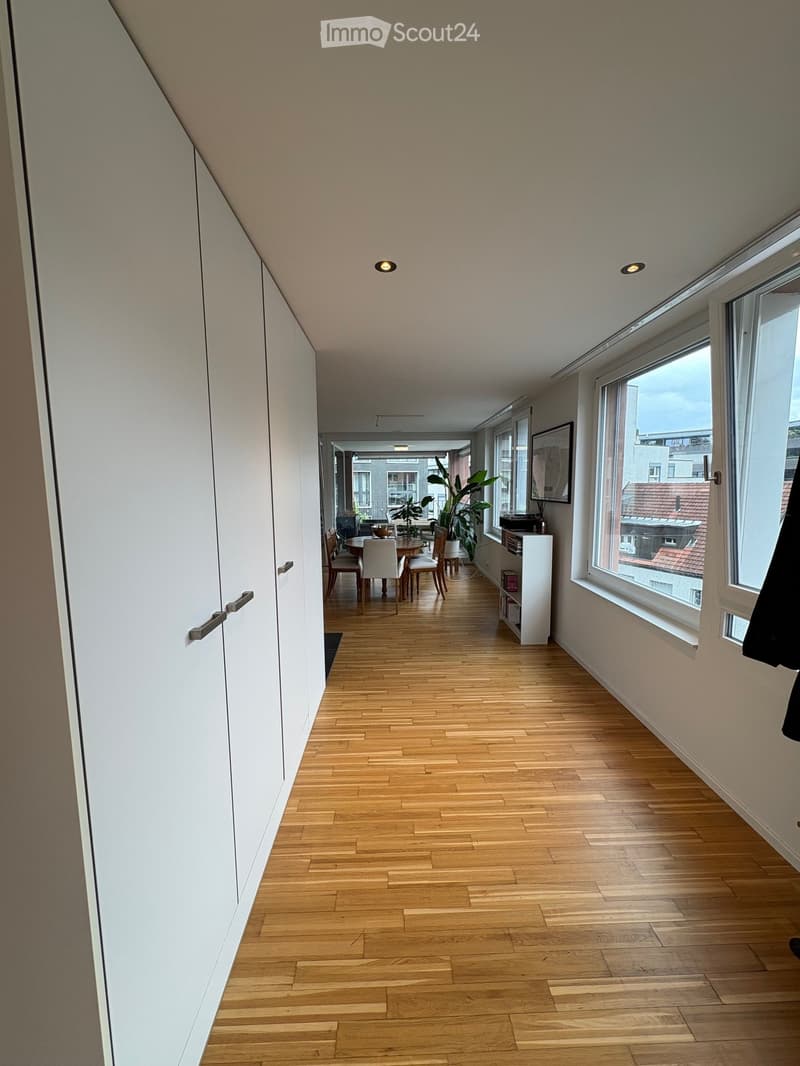 Bright hallway with wooden flooring, white walls, large windows, and recessed lighting. A small dining area is visible at the end with a table and chairs.