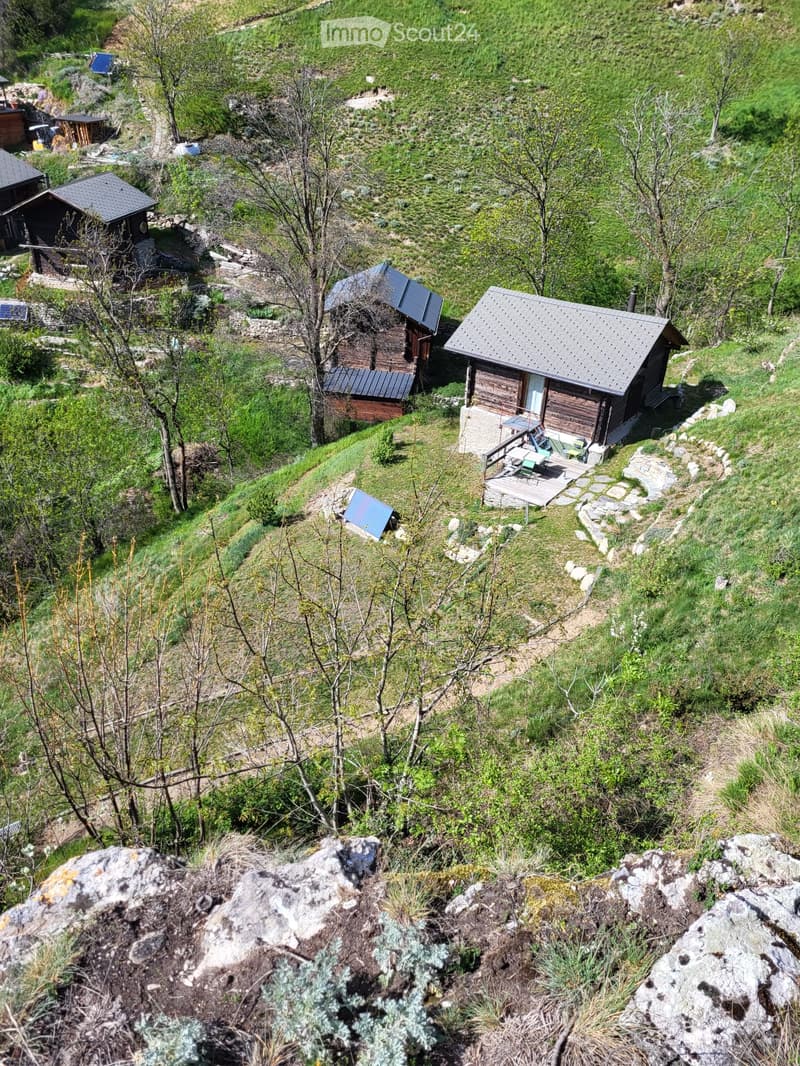 A scenic view of a rural area with several wooden cabins and structures scattered across a grassy hillside, surrounded by trees and natural terrain.