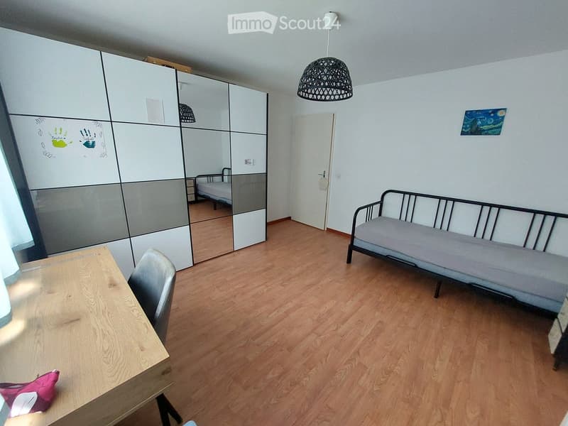 A furnished room with a sofa bed, a wooden table, a chair, a mirror, and a small bed in the background.