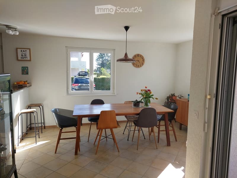 Bright dining area with a wooden dining table surrounded by chairs, two stools, a hanging pendant light, and a potted plant on the table. A large window offers a view of the street and parked cars.