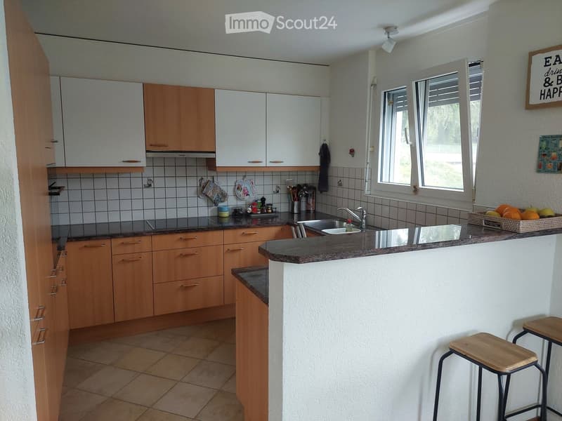 Modern kitchen with light wooden cabinets, granite countertops, and a breakfast bar with two wooden stools. The kitchen features a sink, various small appliances, and a decorative sign on the wall.