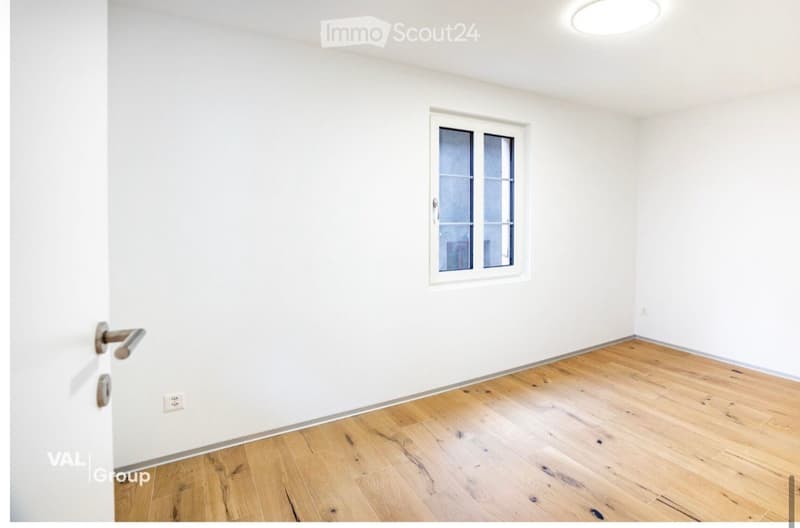 Empty room with white walls, wooden flooring, and a single window with frosted glass panels. The room has a door on the left side and a round ceiling light fixture.