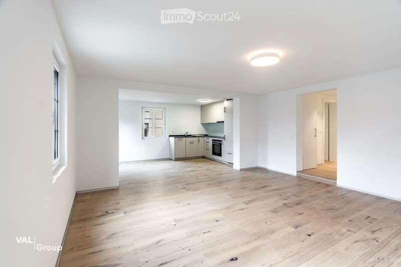 Bright, open-plan living space with light wooden flooring, white walls, and large windows. The kitchen area features modern cabinetry and stainless steel appliances, while the living area is completely empty.