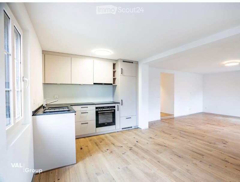 Open-plan space with a modern kitchen area featuring light-colored cabinets, a sink, and an oven, connected to a larger living area with wooden flooring and white walls.