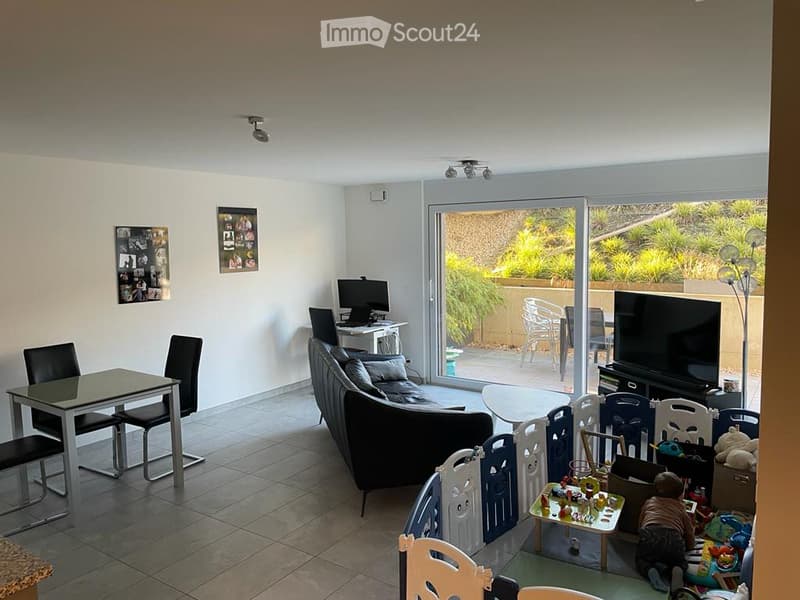A modern living room with a large sliding glass door leading to a balcony. The room features a black sectional sofa, a coffee table, a dining table with chairs, a laptop on a small table, and a TV on a stand. There are also toys and a small table with dri