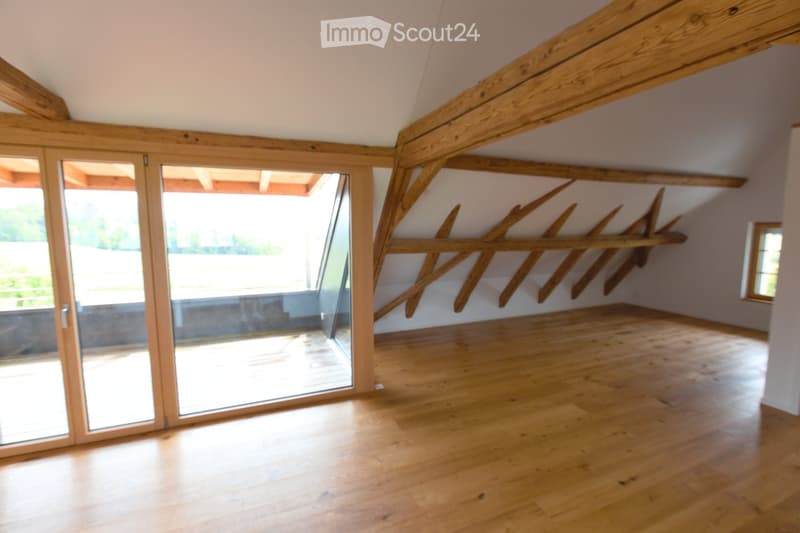 Bright, empty attic room with wooden beams, wooden floor, large glass sliding doors leading to a balcony, and built-in wooden shelves.