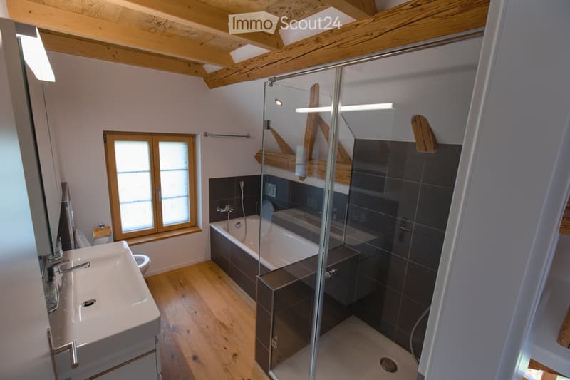 Modern bathroom with wooden flooring, a large window, a sink, a bathtub, a toilet, and a glass-enclosed shower area.