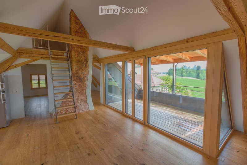 Bright, spacious room with wooden floors, exposed wooden beams, and large glass doors leading to a balcony with scenic views.
