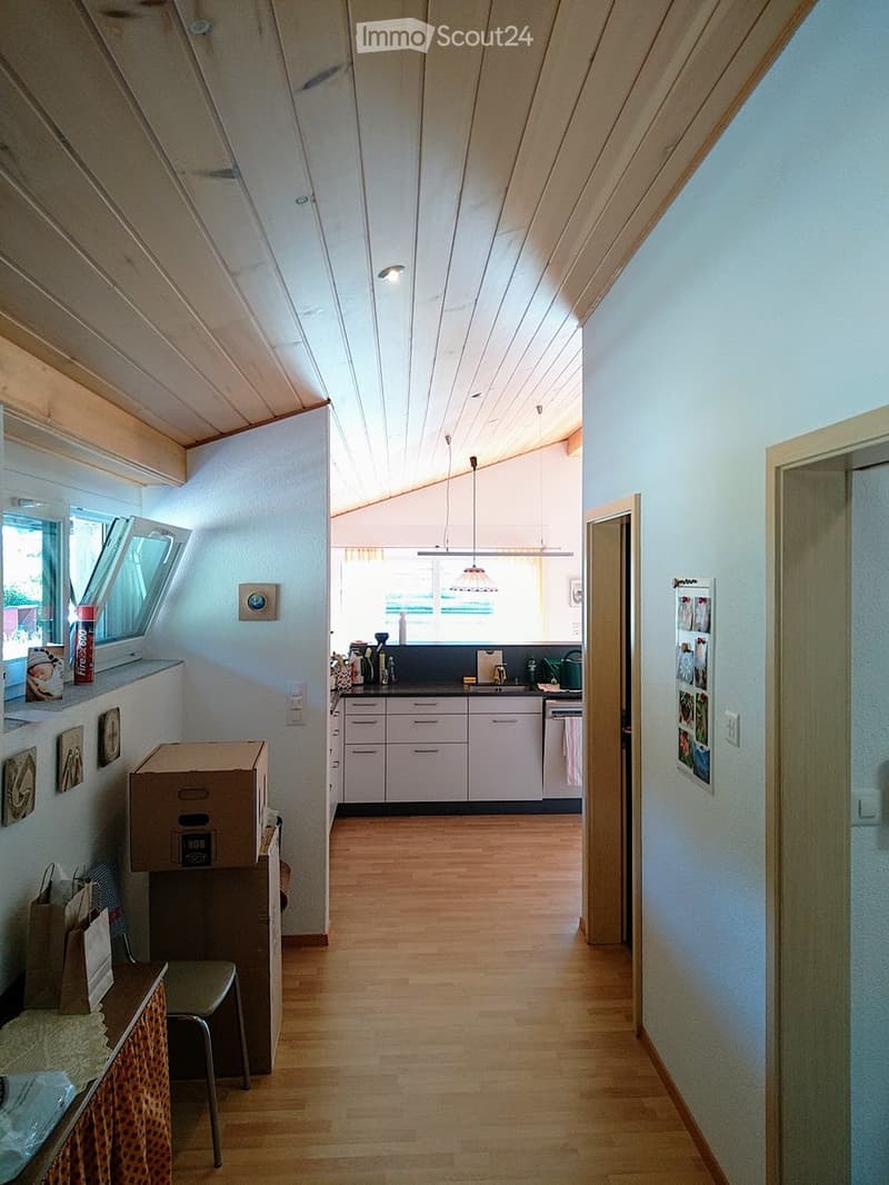 A narrow hallway with wooden flooring, white walls, and a slanted ceiling. The hallway leads to a kitchen area with white cabinets, a sink, and various kitchen items on the counter. There are also some boxes and a small table with a chair in the hallway.