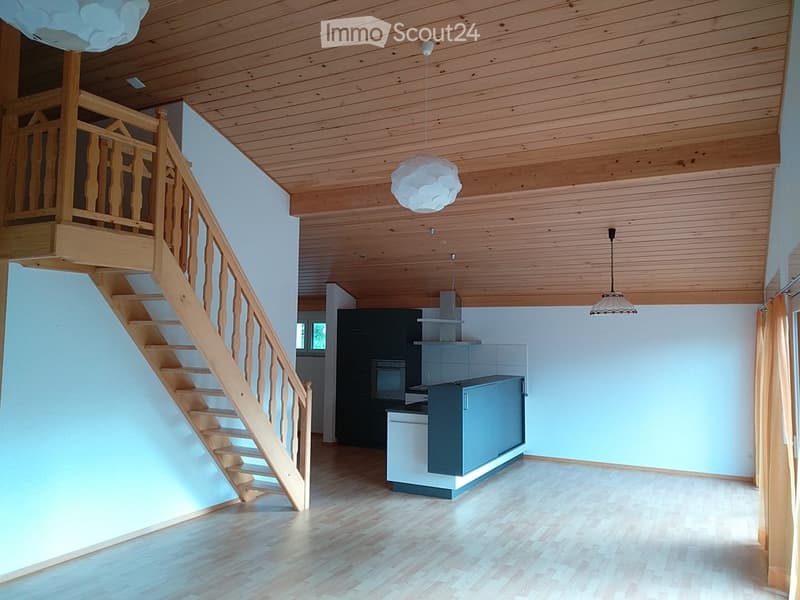 Bright, open-plan living space with wooden flooring, a staircase leading to an upper level, and a modern kitchen area featuring cabinets, a refrigerator, and an oven. The room is illuminated by pendant lights and has a wooden ceiling.