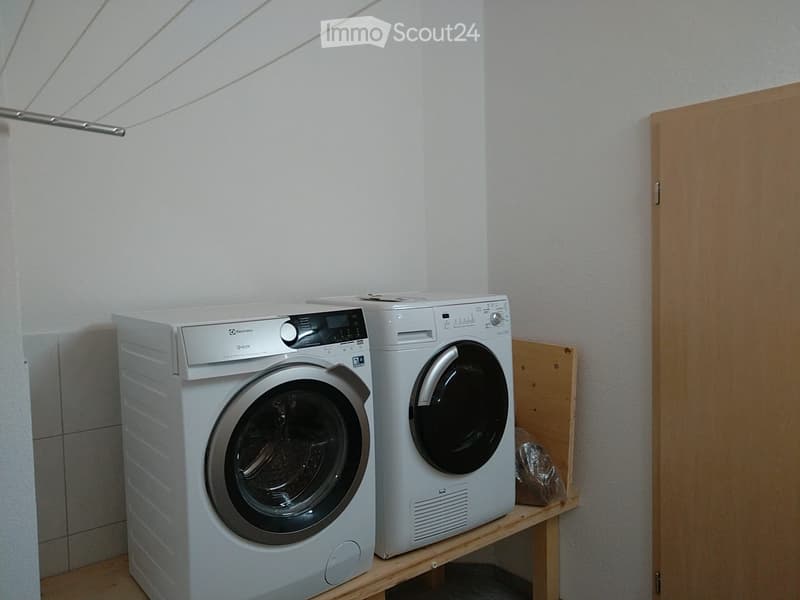 A small room with a washing machine and a dishwasher placed on a wooden shelf. The walls are white, and there is a wooden door on the right side.