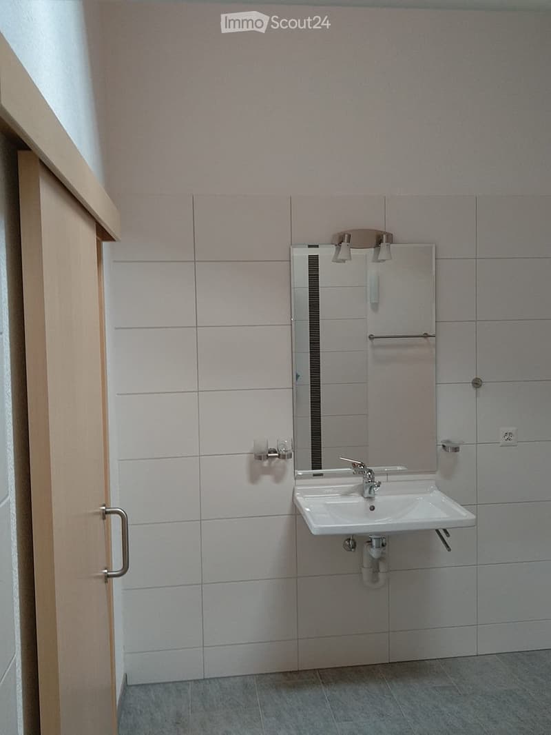 Modern bathroom with a white sink, mirror, and towel rack. The walls are tiled in light gray, and the floor is covered with gray tiles.