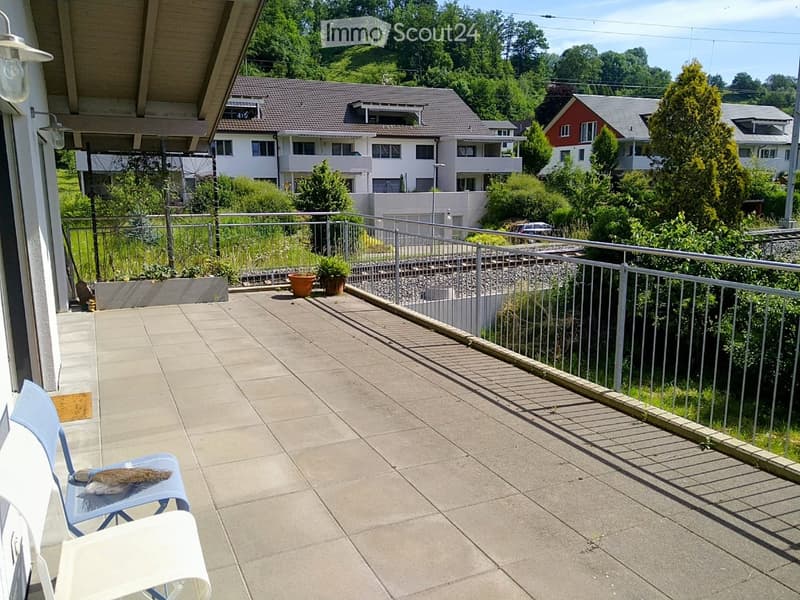 Balcony with concrete floor, metal railing, and a view of the surrounding neighborhood and greenery. There is a blue chair and a white lounge chair on the balcony.