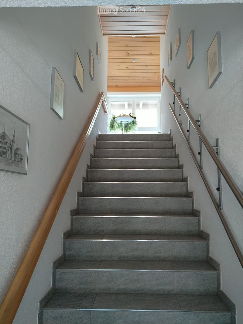 Staircase with grey concrete steps, wooden handrail, and metal railing, leading to a bright landing area with a plant and framed artwork on the walls.