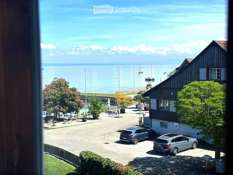 View from a balcony overlooking a parking area, outdoor seating, and a lake with boats. The area includes a wooden building, trees, and a clear blue sky.