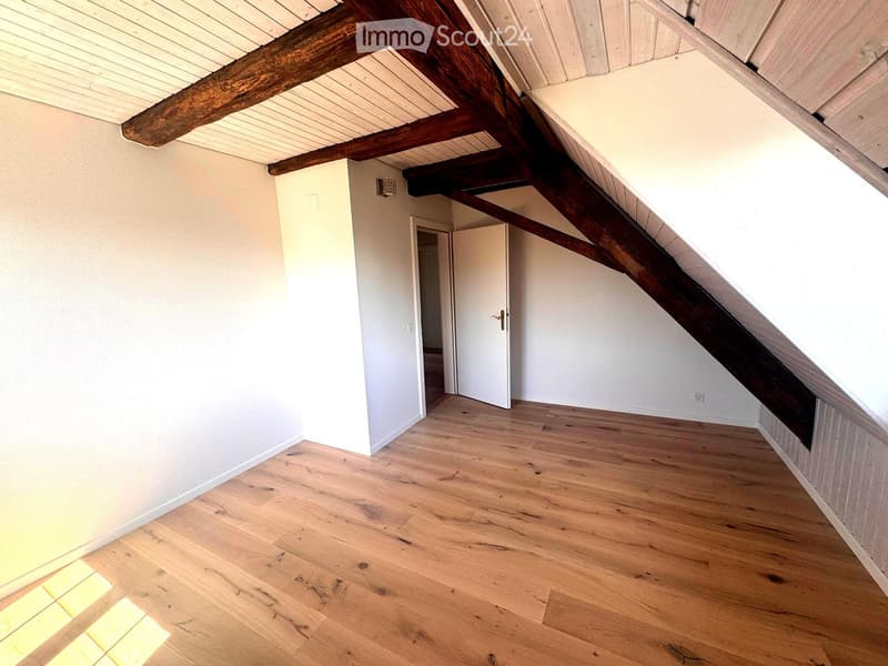 Empty attic room with wooden floor, white walls, exposed wooden beams, and a doorway leading to another space.